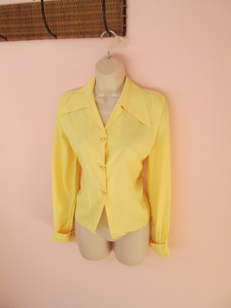 May include: A pale yellow blouse with long sleeves and a pointed collar. The blouse is buttoned down the front with small, round buttons. The cuffs are folded back. The blouse is displayed on a mannequin.