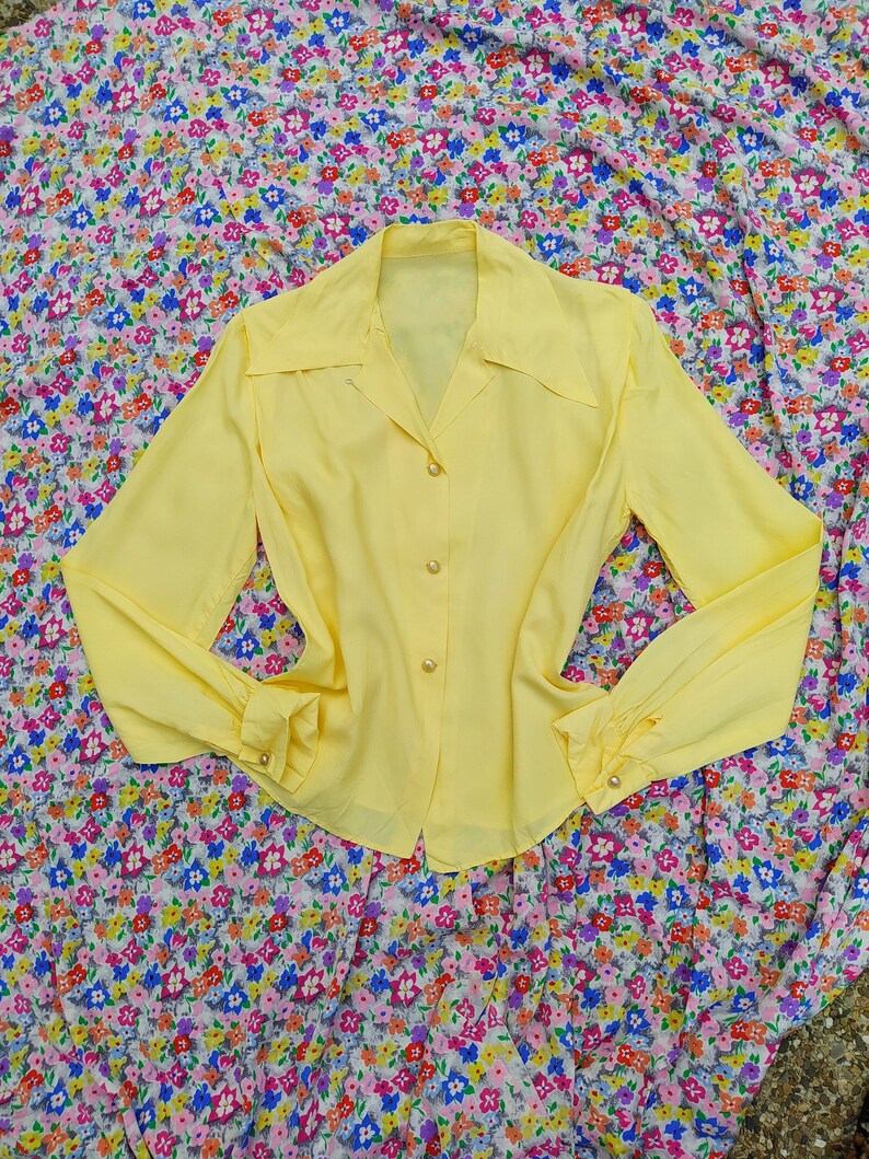 May include: A pale yellow blouse with long sleeves and a collared neckline. The blouse has pearl-like buttons down the front and at the cuffs. The garment is displayed on a floral patterned fabric background.