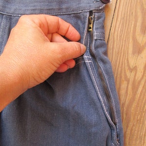 May include: Close-up of denim pants with a side zipper. The pants are a medium blue color with white stitching. A hand is pulling the zipper up. A brown button is visible at the waistband.