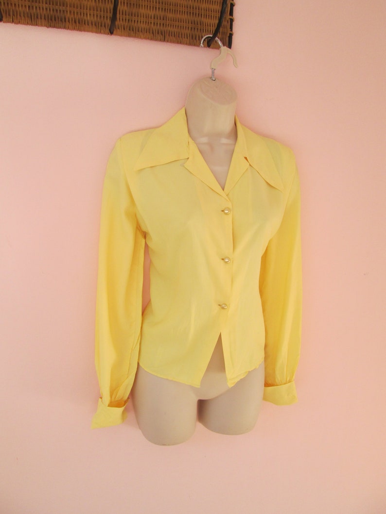 May include: A bright yellow long-sleeve blouse with a collared neckline and button-down front. The blouse has gold-colored buttons and is displayed on a mannequin. The sleeves have buttoned cuffs. The blouse is a solid color.