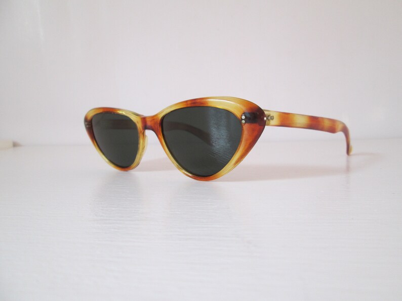 Puede incluir: Gafas de sol de ojo de gato con estampado de carey. Las monturas son una mezcla de marr&oacute;n y amarillo, con lentes oscuras. Las gafas de sol tienen un estilo retro y vintage, sobre un fondo blanco.