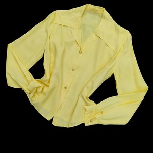 May include: A pale yellow blouse with long sleeves and a collared neckline. The blouse has button closures down the front and at the cuffs. The fabric appears to be a lightweight material.