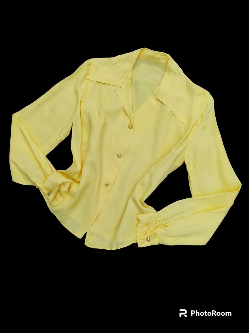 May include: A pale yellow blouse with long sleeves and a collared neckline. The blouse has button closures down the front and at the cuffs. The fabric appears to be a lightweight material.