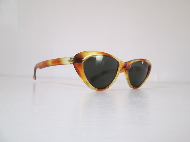 Puede incluir: Gafas de sol vintage de ojo de gato con un patr&oacute;n de carey. Las monturas son &aacute;mbar y marr&oacute;n, con lentes oscuras. Las patillas de las gafas son de un &aacute;mbar m&aacute;s claro.