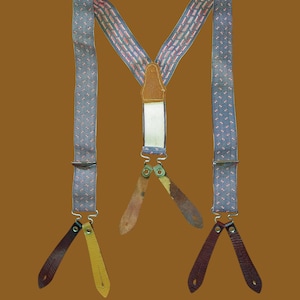 Può includere: Bretelle vintage con un motivo blu e rosso. Le bretelle hanno linguette in pelle marrone, gialla e marrone sbiadito. Sono attaccate a un pezzo di pelle marrone.