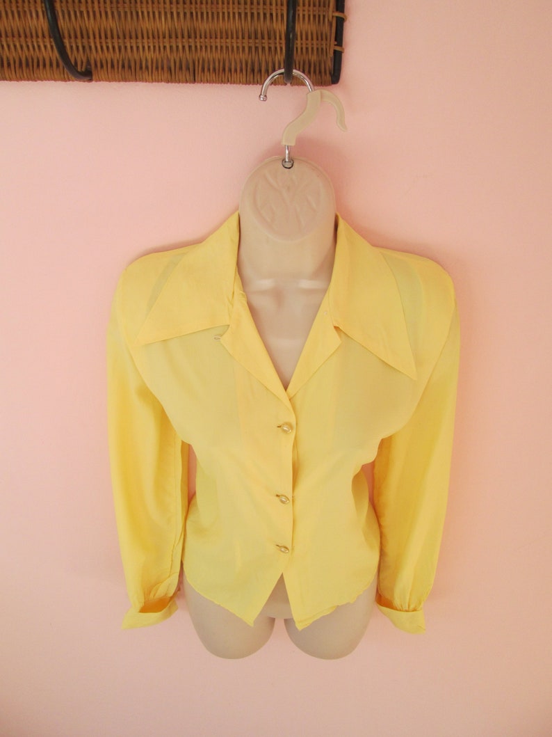 May include: A bright yellow blouse with long sleeves and a collared neckline. The blouse is buttoned down the front and is displayed on a mannequin. The background is a light pink wall.