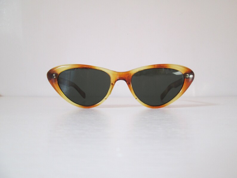 Puede incluir: Gafas de sol de ojo de gato con montura con estampado de carey. Las gafas de sol tienen lentes oscuras y un dise&ntilde;o retro cl&aacute;sico. La montura es una mezcla de tonos &aacute;mbar y marr&oacute;n, con una ligera curva ascendente en los bordes exteriores.