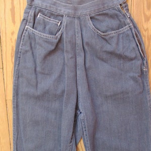 May include: Close-up of blue denim pants with a high waist and a button closure. The pants feature front pockets with white stitching and a zipper detail on the side. The fabric appears to be a medium-wash denim.