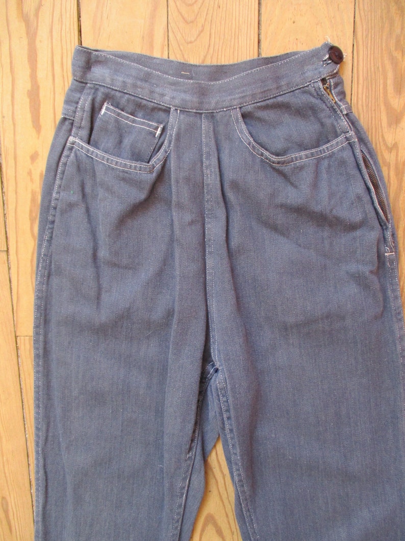 May include: Close-up of blue denim pants with a high waist and a button closure. The pants feature front pockets with white stitching and a zipper detail on the side. The fabric appears to be a medium-wash denim.