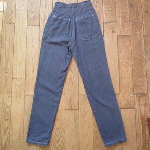 May include: A pair of high-waisted, blue denim pants with two back pockets and white stitching. The pants are laid flat on a wooden floor, showcasing the back view of the garment.