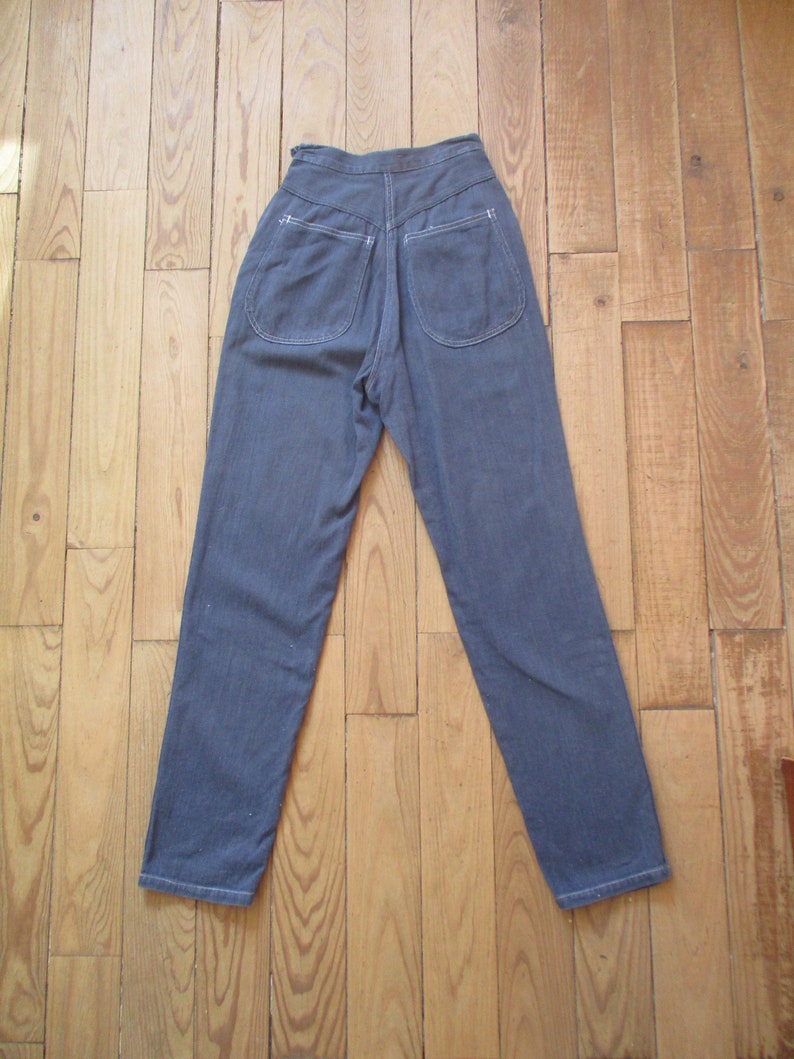 May include: A pair of high-waisted, blue denim pants with two back pockets and white stitching. The pants are laid flat on a wooden floor, showcasing the back view of the garment.