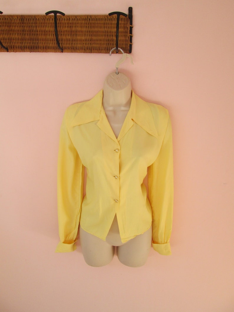 May include: A pale yellow long-sleeved blouse with a collared neckline and button-down front. The blouse is displayed on a mannequin and has buttoned cuffs. The background is a light pink wall.