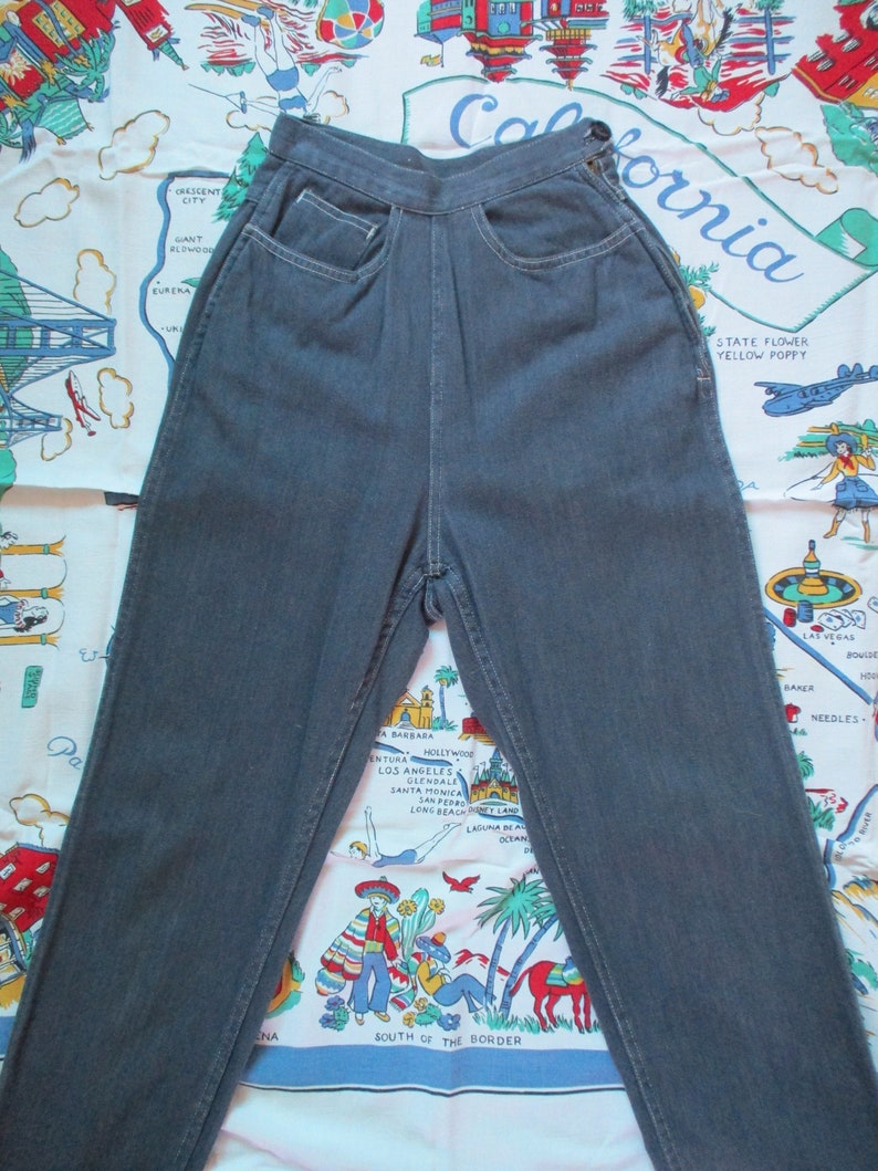 May include: Dark gray denim jeans with a high waist and classic five-pocket design. The jeans have a button and zipper closure and are made of a sturdy denim material. The jeans are displayed on a colorful California-themed fabric.