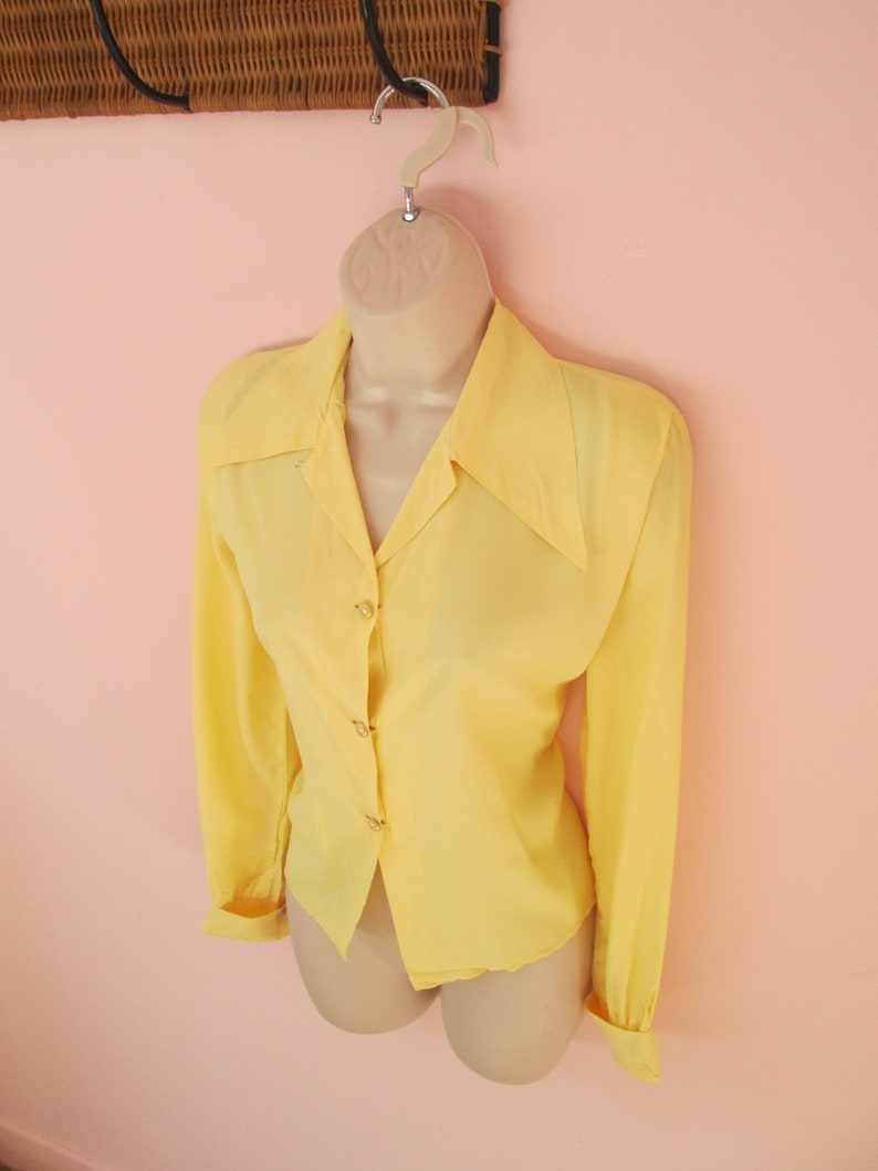 May include: A pale yellow long-sleeved blouse with a classic collar and button-down front. The blouse is displayed on a mannequin, showcasing its tailored fit and design. The cuffs are buttoned, and the fabric appears to be lightweight.