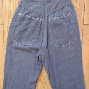 May include: A pair of high-waisted, blue denim pants. The pants feature two rounded back pockets with white stitching and a V-shaped yoke. The denim has a slightly faded appearance, and the pants are displayed on a wooden surface.