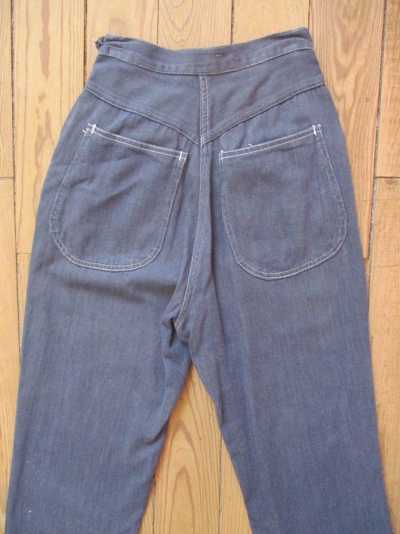 May include: A pair of high-waisted, blue denim pants. The pants feature two rounded back pockets with white stitching and a V-shaped yoke. The denim has a slightly faded appearance, and the pants are displayed on a wooden surface.