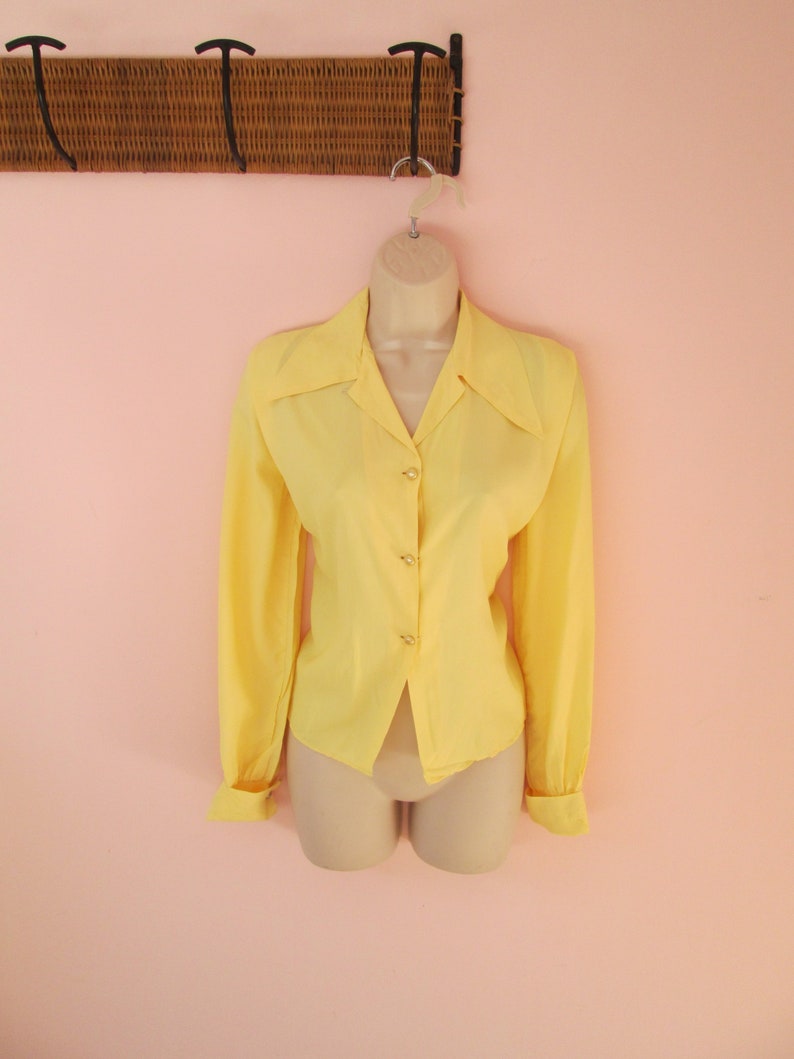 May include: A vintage yellow blouse with long sleeves and button details. The blouse is displayed on a mannequin against a pink background. The blouse has a collared neckline and buttoned cuffs.