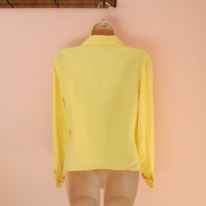 May include: A bright yellow long-sleeved blouse with a classic collar and buttoned cuffs. The blouse is displayed on a mannequin, showcasing its shape and design. The garment hangs from a white hanger against a soft pink background.