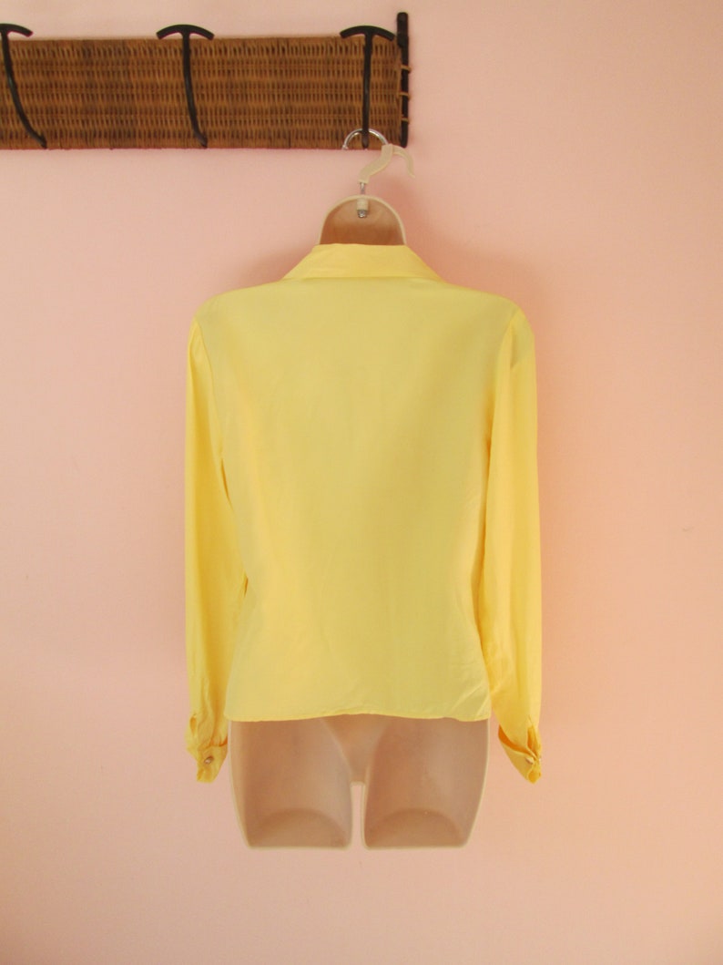 May include: A bright yellow long-sleeved blouse with a classic collar and buttoned cuffs. The blouse is displayed on a mannequin, showcasing its shape and design. The garment hangs from a white hanger against a soft pink background.
