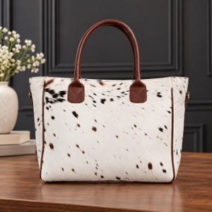 May include: A tote bag featuring a white and brown cowhide pattern with a textured hair-on-hide finish. The bag includes two brown leather handles, brown leather trim along the side seams, and gold-toned metal hardware attachments on the sides.