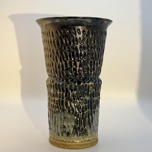 May include: A tall ceramic vase with a flared rim and a textured, hammered surface. The exterior features a dark, glossy black glaze that transitions into a lighter, speckled beige tone at the base, highlighting the deep, vertical indentations.