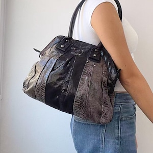 Puede incluir: Un bolso de hombro con un exterior de patrón de piel de serpiente en gris oscuro y negro con paneles verticales. El bolso incluye correas de aspecto de cuero negro, herrajes de metal plateado y una placa de logo metálica rectangular.