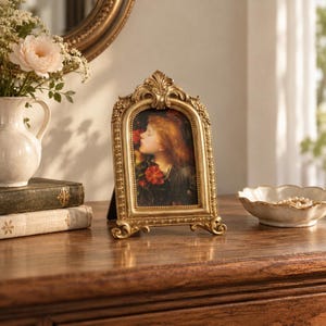 Puede incluir: Un marco de fotos dorado, arqueado y ornamentado, con una parte superior de volutas decorativas y bordes con cuentas. El marco contiene el retrato de una persona con cabello rojizo y piel clara, rodeada de flores rojas. Se apoya sobre una superficie de madera.