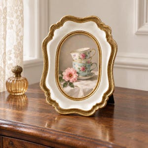 May include: An oval picture frame with a white center and a gold scalloped border. The frame contains an image of two stacked floral teacups, a saucer, and a pink flower on a white cloth. The frame has a stand on the back.