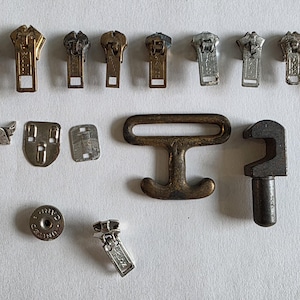 May include: A collection of assorted metal hardware components including nine zipper sliders in various brass and silver tones, three small metal fasteners, a large bronze-toned T-shaped buckle, a dark metal hook, and a circular metal button.