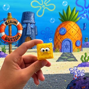 Pode incluir: Uma pequena figura quadrada e amarela do personagem Bob Esponja, com grandes olhos brancos, um nariz pequeno e uma boca sorridente, apresentando uma textura porosa na superfície, segurada contra um fundo subaquático colorido com uma casa abacaxi.