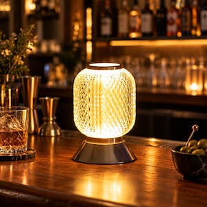 May include: A cylindrical table lamp with a textured, diamond-patterned glass shade that emits a warm, golden glow. The lamp features a smooth, metallic base and sits on a wooden surface, providing illumination through its intricate glass structure.