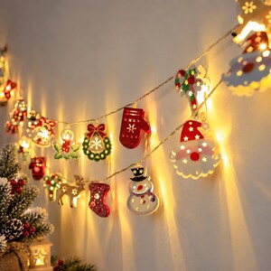May include: A string of LED Christmas lights featuring various wooden ornaments including a Santa Claus face, a snowman, a red mitten, a stocking, a reindeer, a wreath, and a candy cane, all emitting a warm yellow glow against a plain wall.