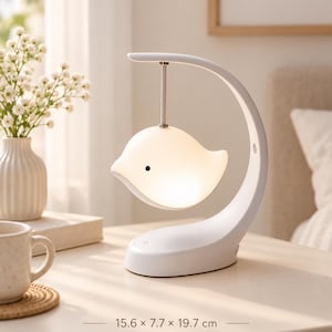 Può includere: Una lampada bianca a forma di uccellino sospesa a un braccio curvo su una base. La lampada emette una luce soffusa e presenta un piccolo occhio nero. Il testo stampato sulla superficie indica dimensioni di 15,6 x 7,7 x 19,7 cm. La struttura è liscia e opaca.