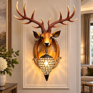 May include: A wall-mounted light fitting featuring a three-dimensional deer head sculpture with large, brown antlers. The deer is set within an oval frame, and a teardrop-shaped lamp with a faceted crystal-like cage hangs from its neck.