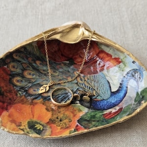 May include: A decorative seashell trinket dish with a gold-painted rim, featuring a colorful decoupage design of a peacock and orange flowers. Inside the shell rests a thin gold chain necklace with a leaf charm, a gold ring, and stud earrings.