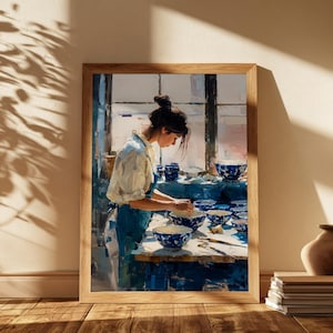 May include: A framed wall art print depicting a person in a white shirt and blue apron working with blue and white patterned ceramic bowls on a table in a sunlit studio. The scene features multiple bowls arranged on the workspace near a window.