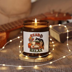 Pode incluir: Uma vela acesa num frasco de vidro castanho com um rótulo branco com um gato preto e branco sobre livros, uma caneca, uma abóbora e folhas de outono, com o texto READ & RELAX impresso a vermelho e verde na frente do rótulo.
