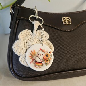 May include: A keychain featuring a round white disc with a colorful illustration of a small bird holding a bouquet of sunflowers and wearing a German flag patterned apron, attached to a cream-colored crocheted flower shape.