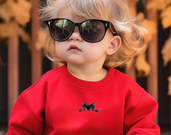 May include: A child wearing black sunglasses and a red sweatshirt. The sweatshirt features a small graphic of a black cat peeking over a line. The background is blurred with fall foliage.