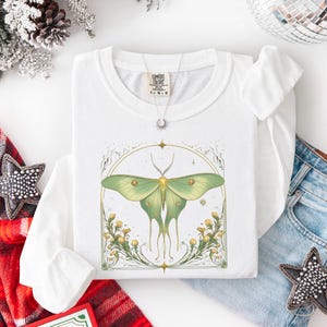 May include: White long-sleeved shirt featuring a large green and yellow moth design within a decorative frame. The shirt has a crew neck and the brand name "Comfort Colors" is visible. A silver necklace with a sun pendant is also present.