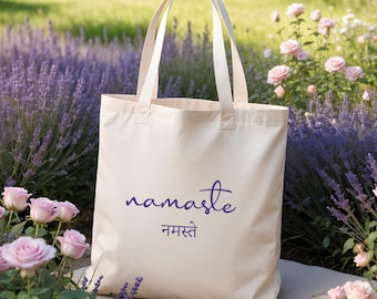 May include: Cream-colored tote bag with the word "namaste" in a dark blue script, along with the Hindi translation. The bag has two long handles and is set against a backdrop of lavender and pink roses.