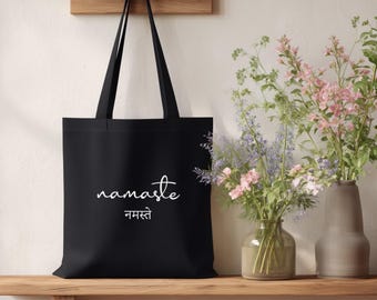 May include: Black tote bag with the word "namaste" in white script and Hindi text. The bag hangs from a wooden rack on a light-colored wall. A wooden shelf holds the bag and two vases with flowers.