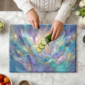 May include: A rectangular cutting board with a stained glass design in shades of blue, purple, and gold. A person is slicing a cucumber on the board with a knife. The board has a decorative, abstract floral pattern.