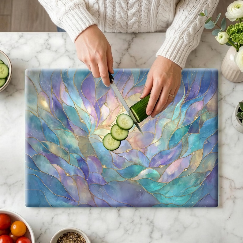 May include: A rectangular cutting board with a stained glass design in shades of blue, purple, and gold. A person is slicing a cucumber on the board with a knife. The board has a decorative, abstract floral pattern.