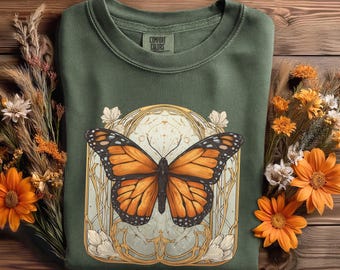 May include: Green sweatshirt with a large orange and black butterfly graphic. The butterfly is centered within a decorative gold and cream-colored frame. The sweatshirt has a crew neck and the brand name "Comfort Colors" is visible on a tag.