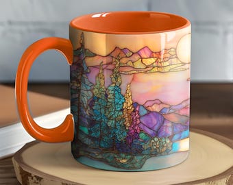 Könnte beinhalten: Eine Keramik-Tasse mit orangefarbenem Henkel und Innenraum. Die Außenseite zeigt eine farbenfrohe Landschaft im Glasmalerei-Stil mit Bäumen, Bergen und einer Sonne. Die Tasse steht auf einem Holzuntersetzer.