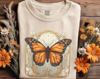 May include: Cream-colored sweatshirt featuring a large, detailed butterfly design. The butterfly has orange and black wings, set within an ornate, gold-toned frame. The sweatshirt is made by Comfort Colors and is surrounded by dried flowers.