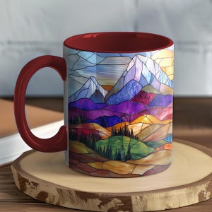 May include: A ceramic mug with a burgundy handle and interior. The exterior features a colorful stained-glass design of a mountain landscape with purple, blue, orange, and green hues. The mug sits on a wooden coaster.