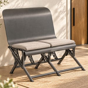 Puede incluir: Un banco de metal gris oscuro con un respaldo curvo y dos cojines de asiento rectangulares de color gris claro separados. El banco está sostenido por una compleja estructura de marco plegable geométrica con patas anguladas y barras.