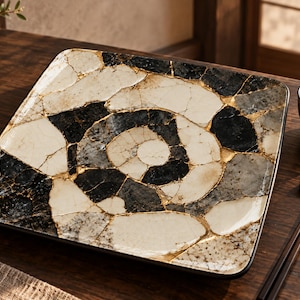 Può includere: Un piatto quadrato in ceramica con un motivo a mosaico composto da segmenti neri, bianchi e grigi dalla consistenza simile alla pietra, uniti da sottili linee color oro metallizzato. Il design si irradia dal centro in una spirale.